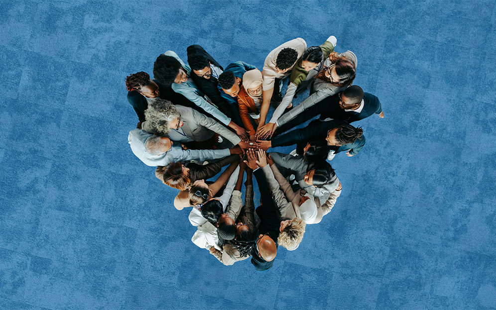 Eine Gruppe von Menschen verschiedener Herkunft hält sich an den Händen. Das Bild ist von oben aufgenommen und zeigt aus dieser Perspektive, dass die Menschen ein Herz bilden.