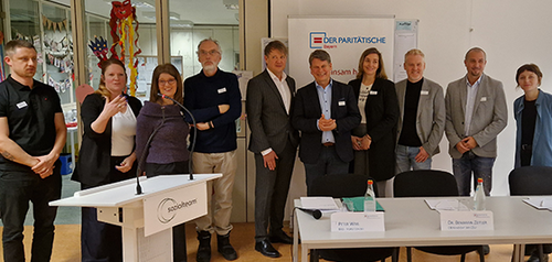 Neun Personen stehen in einem Konferenzraum hinter einem Rednerpult und einem Tisch mit Namensschildern.